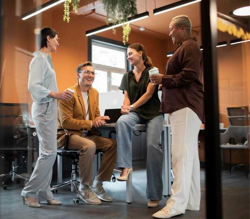 Colegas de trabalho diversos reúnem-se em um escritório moderno, sorrindo e conversando enquanto tomam café, criando uma atmosfera acolhedora, colaborativa e receptiva.