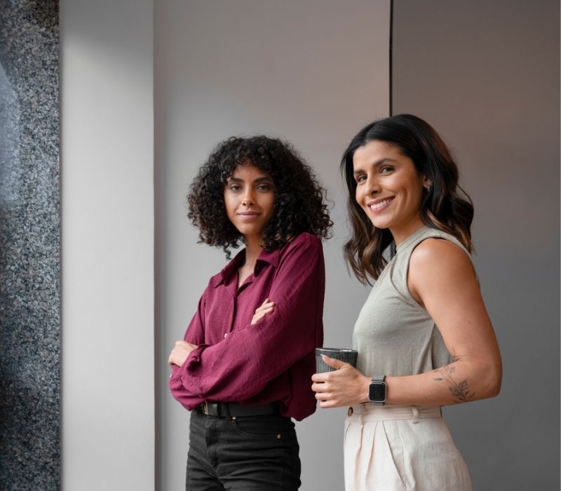 Duas mulheres posam em um ambiente interno: uma de cabelos cacheados e braços cruzados, a outra sorrindo enquanto segura uma xícara. Retrato profissional moderno e simpático.